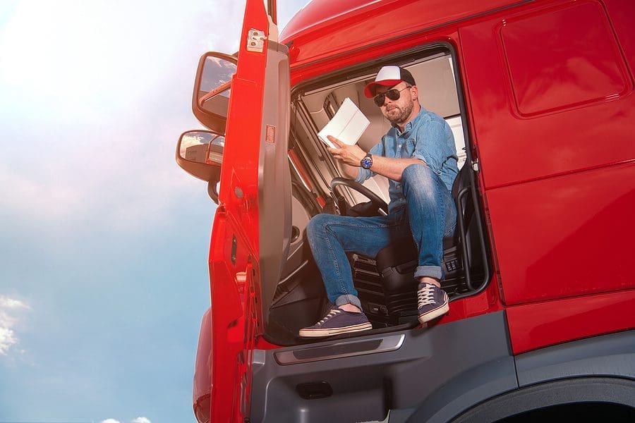 Caucasian Lorry Driver Sitting in the Cabin of His Red Semi Truck Carefully Checking Transit Papers Before Starting a Route With a Large Cargo Delivery. Heavy Duty Transportation Theme.
