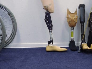 Four different types of prosthetic legs are lined up against a wall next to a wheelchair